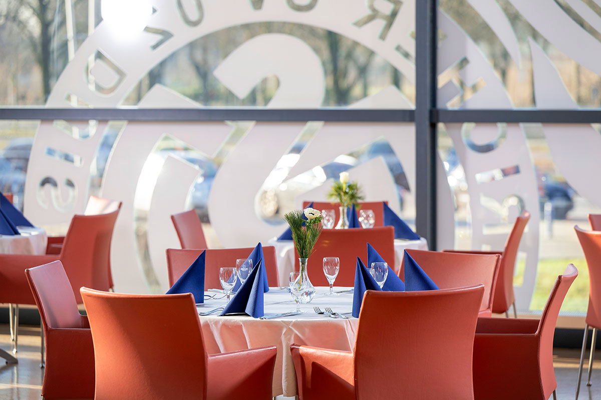 Eingedeckte Tische mit roten Stühlen, im Hintergrund das "MSV Duisburg"-Logo auf einer großen Fensterscheibe.