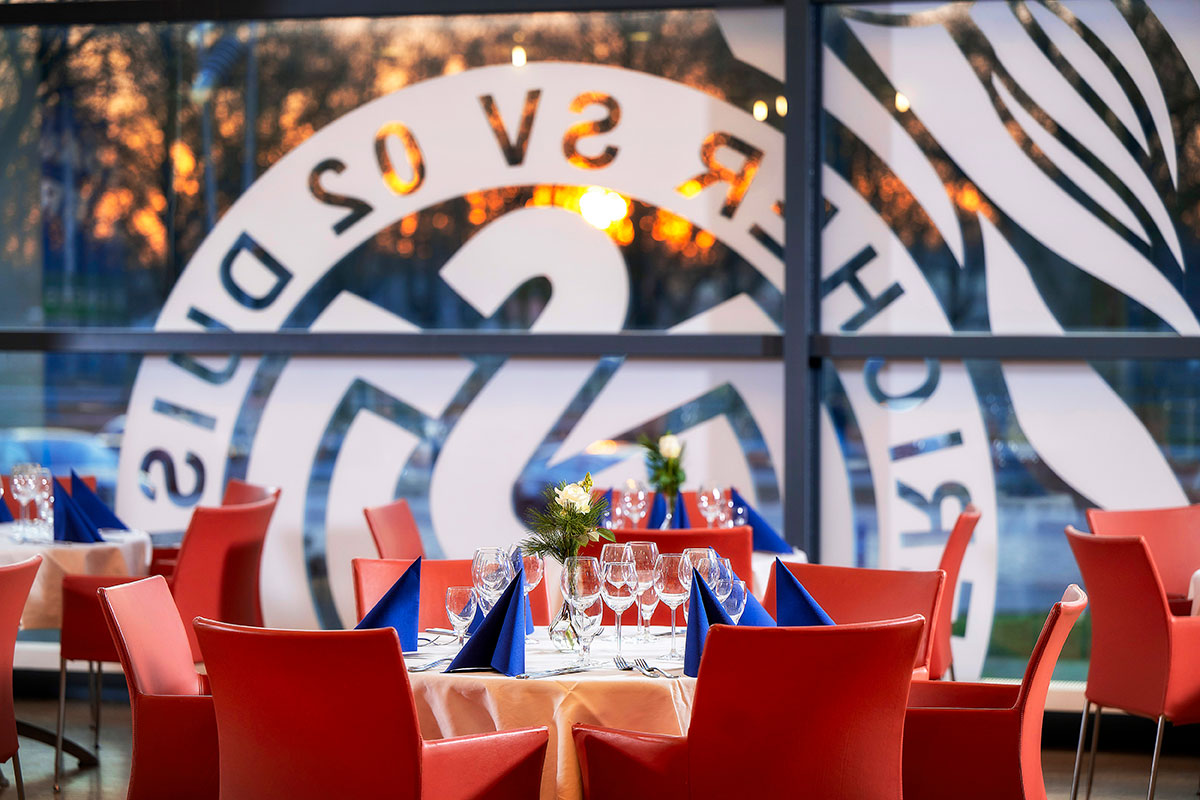 Eingedeckte Tische mit roten Stühlen, im Hintergrund das "MSV Duisburg"-Logo auf einer großen Fensterscheibe.