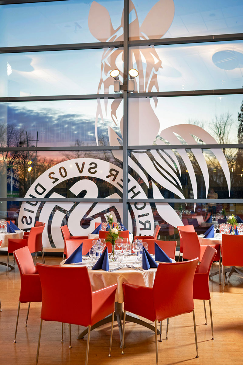 Die Business Lounge mit runden Tischen, eingedeckt mit weißen Tischdecken, roten Stühlen und blauen Servietten.