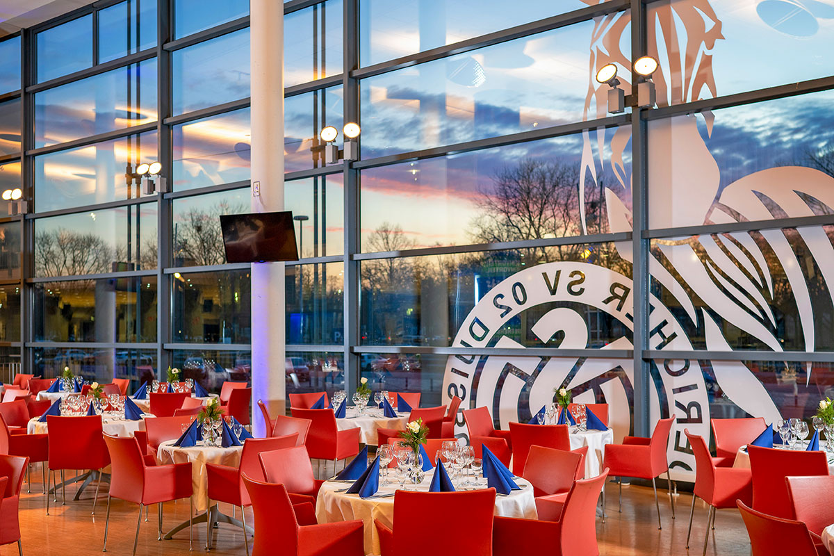 Viele eingedeckte Tische mit roten Stühlen, im Hintergrund das "MSV Duisburg"-Logo auf einer großen Fensterscheibe.