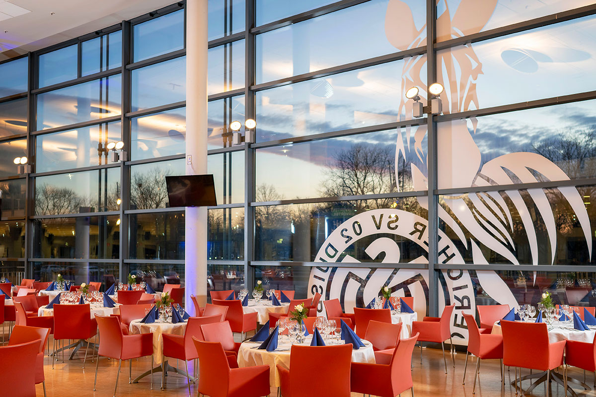 Viele eingedeckte Tische mit roten Stühlen, im Hintergrund das "MSV Duisburg"-Logo auf einer großen Fensterscheibe.