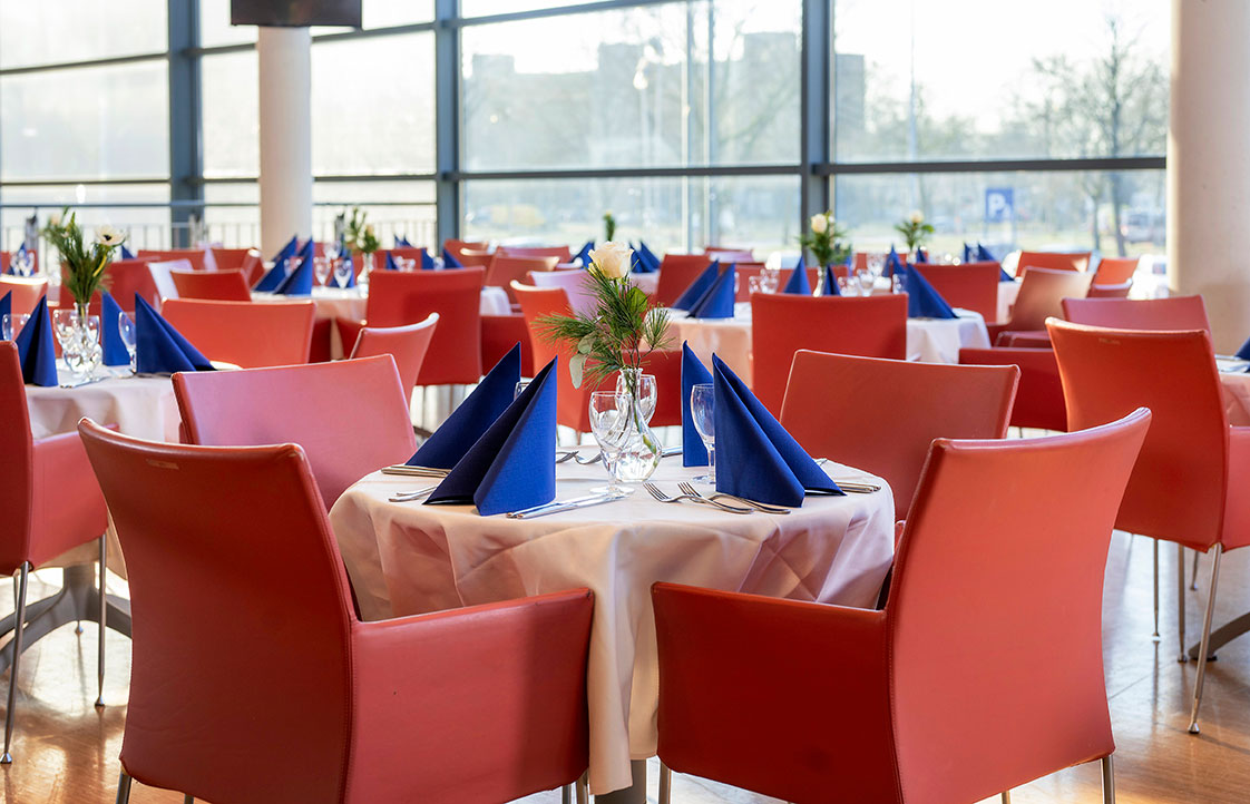 Eingedeckte Tische mit roten Stühlen, im Hintergrund eine große Fensterfront.