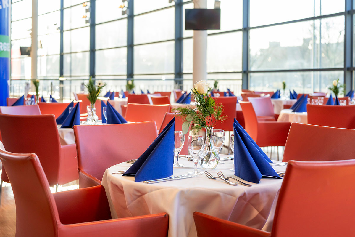 Eingedeckte Tische mit roten Stühlen, im Hintergrund eine große Fensterfront.