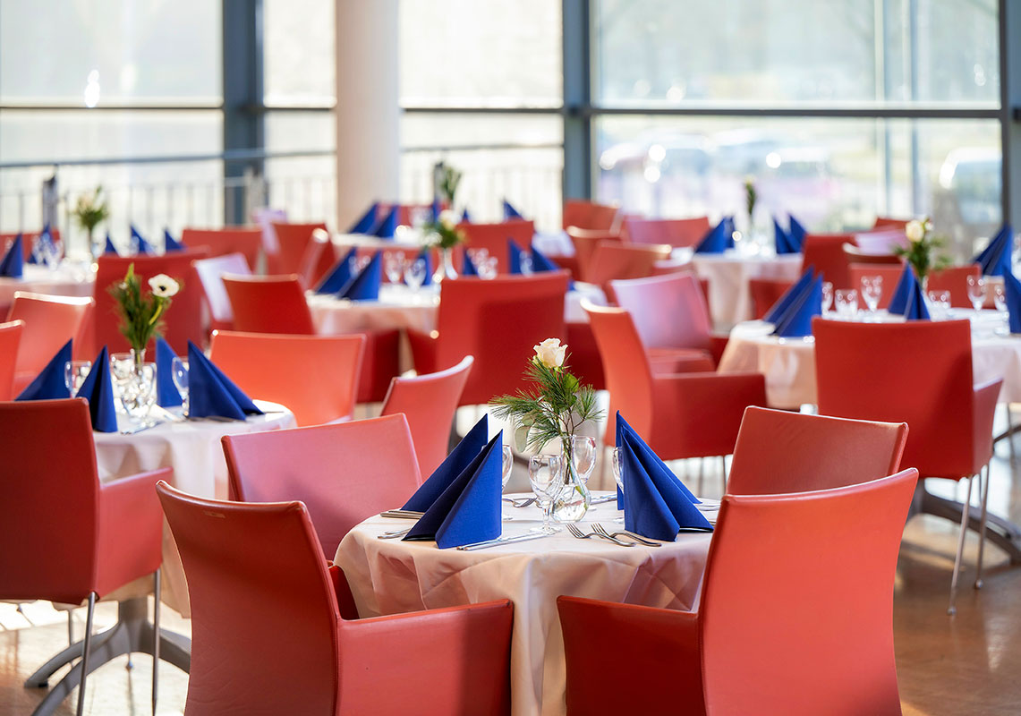 Die Business Lounge mit runden Tischen, eingedeckt mit weißen Tischdecken, roten Stühlen und blauen Servietten.