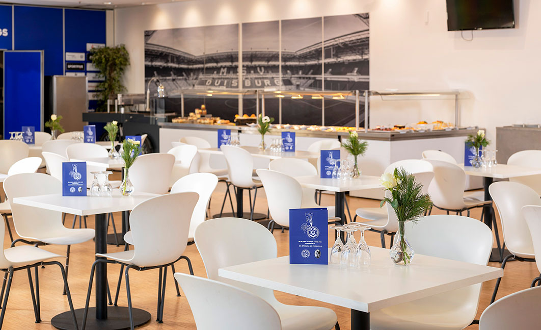 Eine Lounge mit weißen Tischen und Stühlen, davor ein Buffet und ein Stadionwandbild.