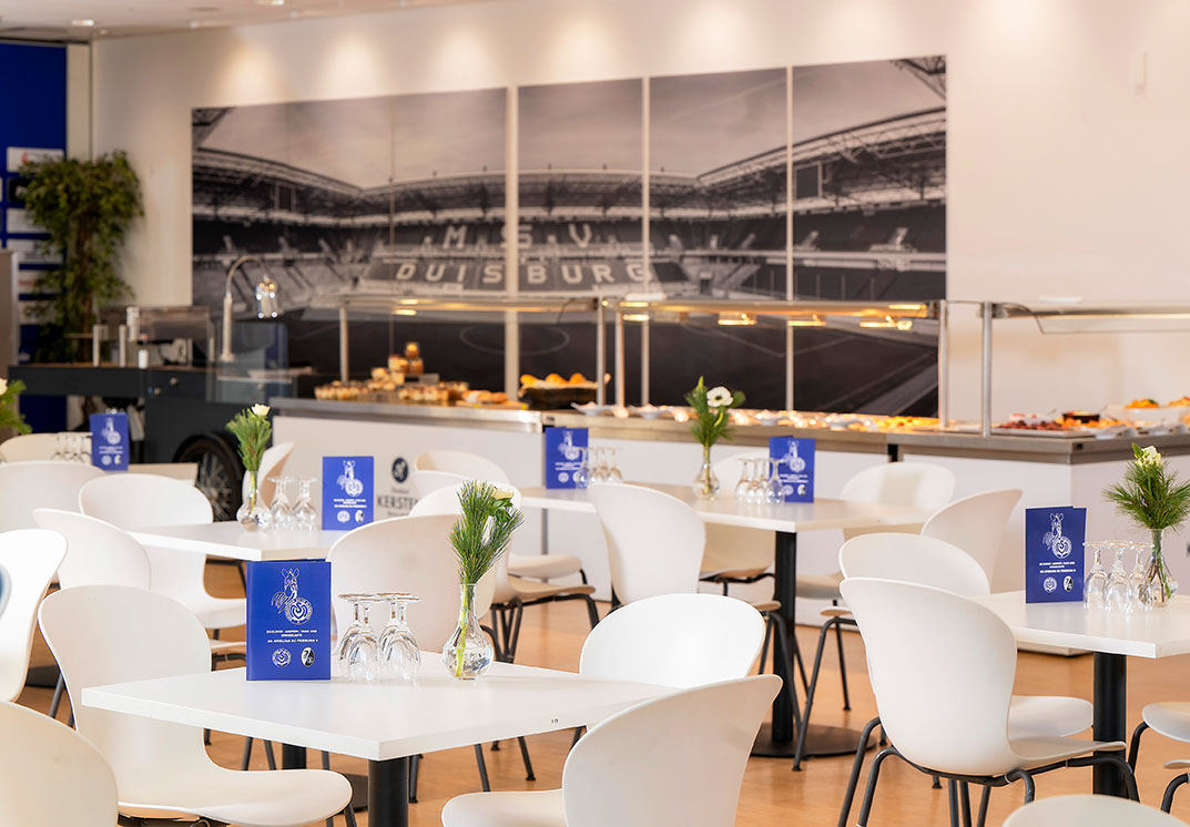 Eine Lounge mit weißen Tischen und Stühlen, davor ein Buffet und ein Stadionwandbild.