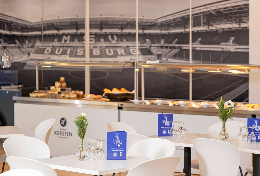 Eine Lounge mit weißen Tischen und Stühlen, davor ein Buffet und ein Stadionwandbild.