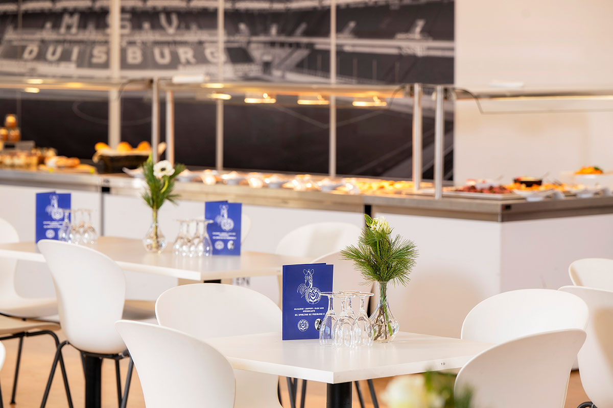 Eine Lounge mit weißen Tischen und Stühlen, davor ein Buffet und ein Stadionwandbild.