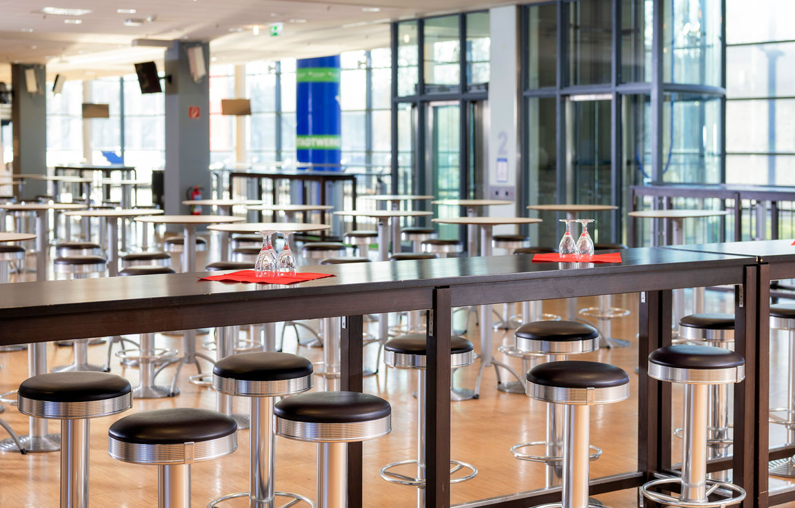 Eine großer heller Eventbereich mit vielen Stehtischen und Barhockern. Dahinter eine große Fensterfront.