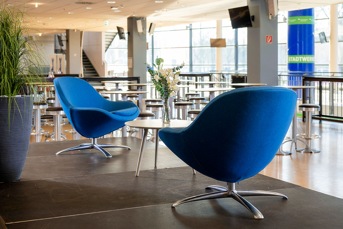 Zwei blaue Sessel stehen um einen kleinen Tisch in einem großen Raum mit vielen weiteren Tischen und Stühlen im Hintergrund.