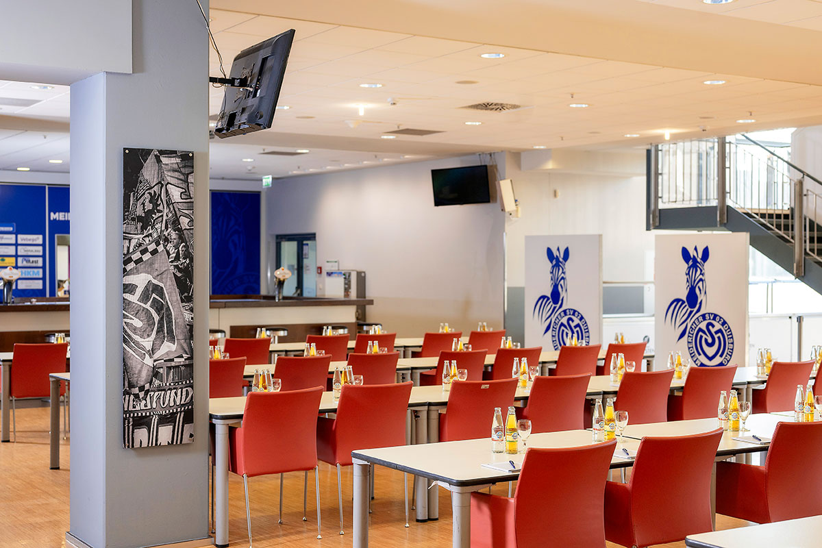 Ein großer Veranstaltungsraum mit Tischreihen und roten Stühlen. Im Hintergrund stehen MSV-Zebralogos