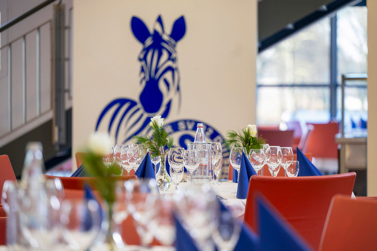 Ein festlich gedeckter Tisch mit Gläsern, blauen Servietten und einem Zebra-Logo im Hintergrund.