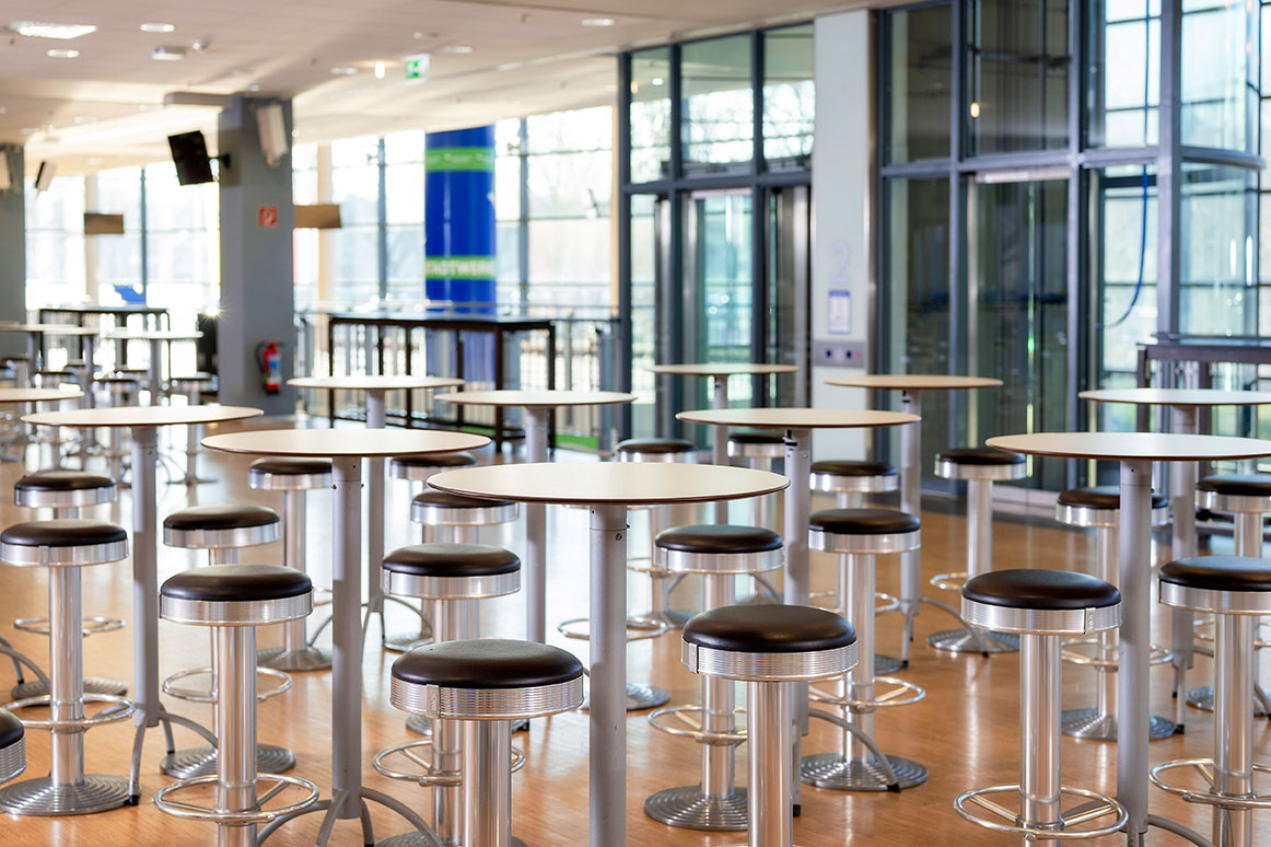 Eine großer heller Eventbereich mit vielen Stehtischen und Barhockern. Dahinter eine große Fensterfront.