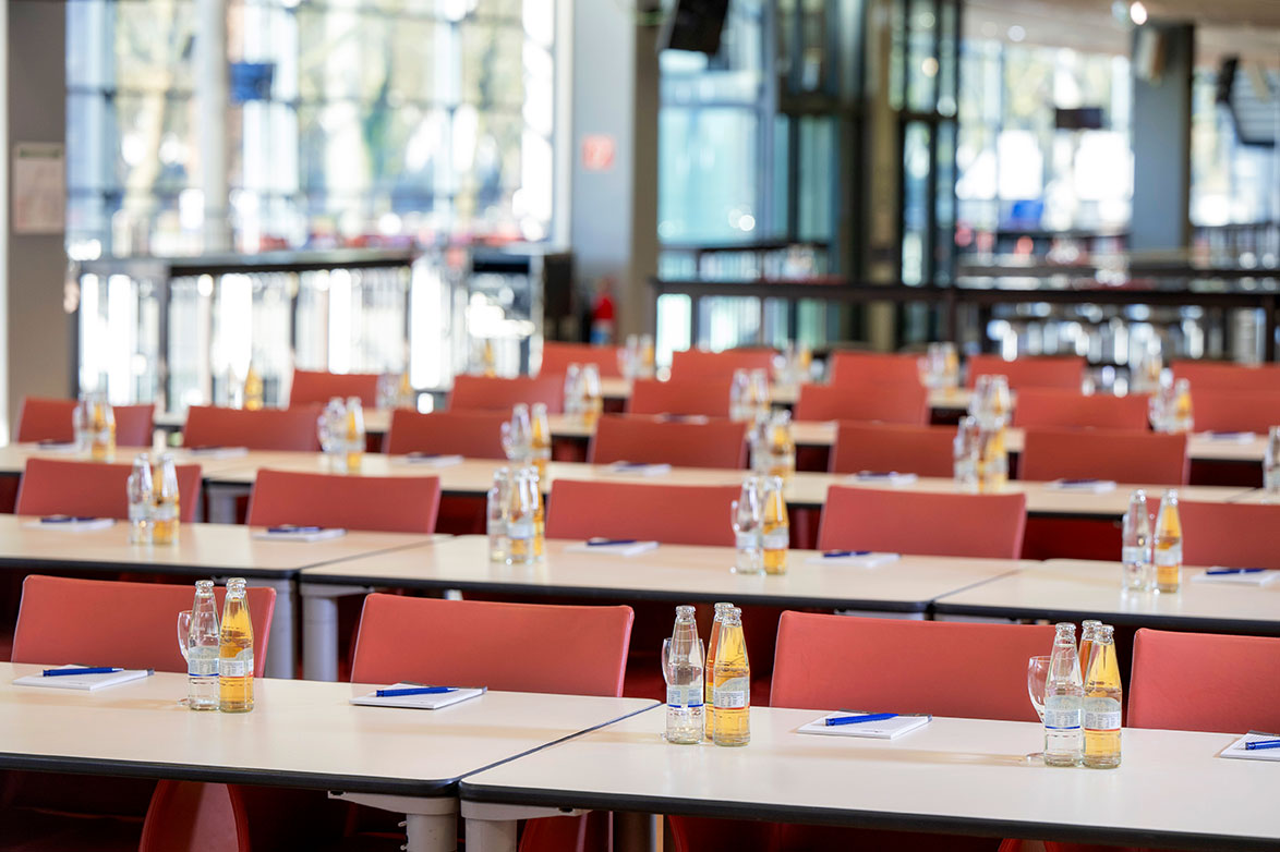 Eine großer Eventbereich mit vielen hintereinander aufgereihten Stühlen und weißen Tischen mit Getränken und Notizblöcken.