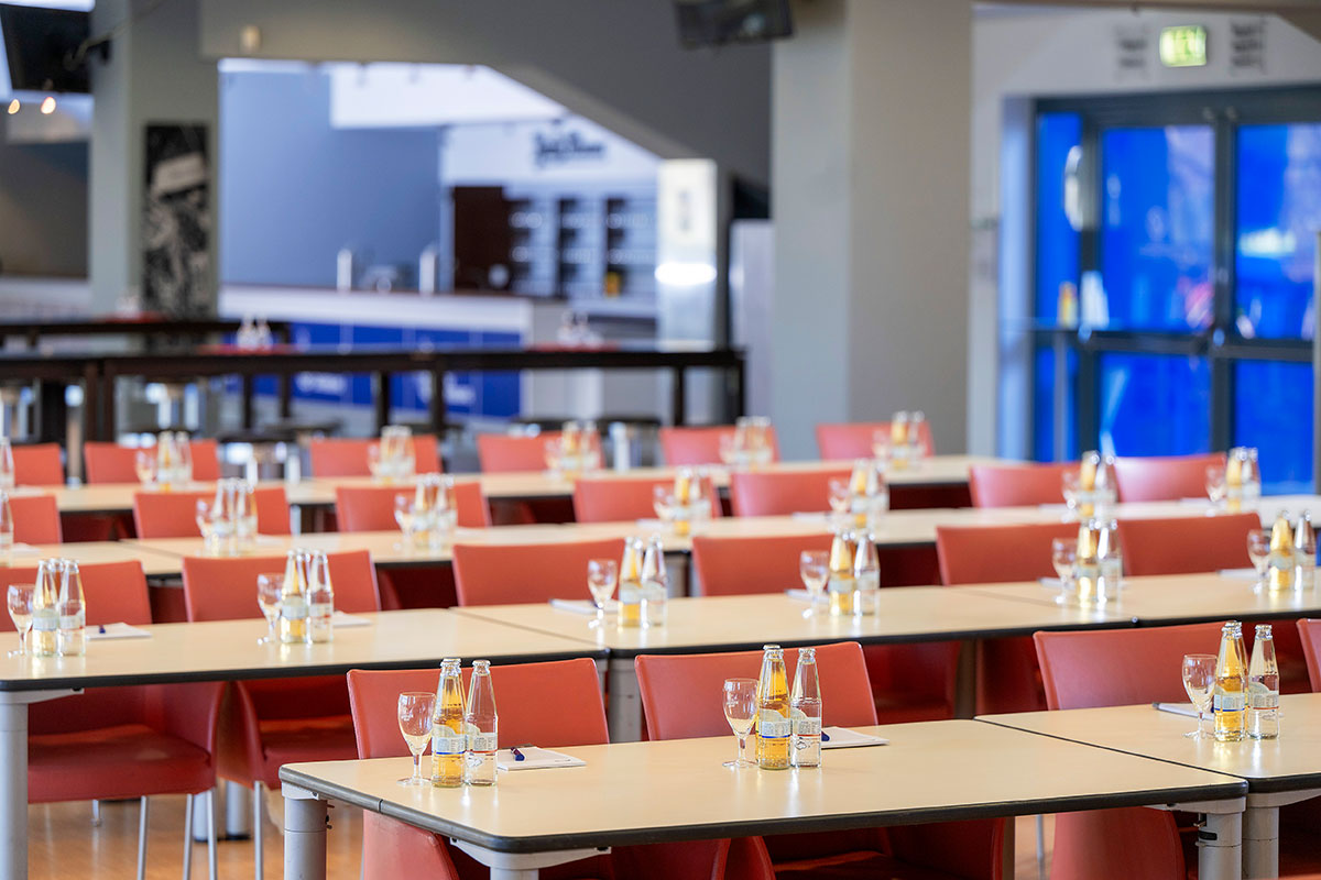 Ein großer Veranstaltungsraum mit Tischreihen und roten Stühlen. Die Tische sind mit Getränken und Notizblöcken eingedeckt.