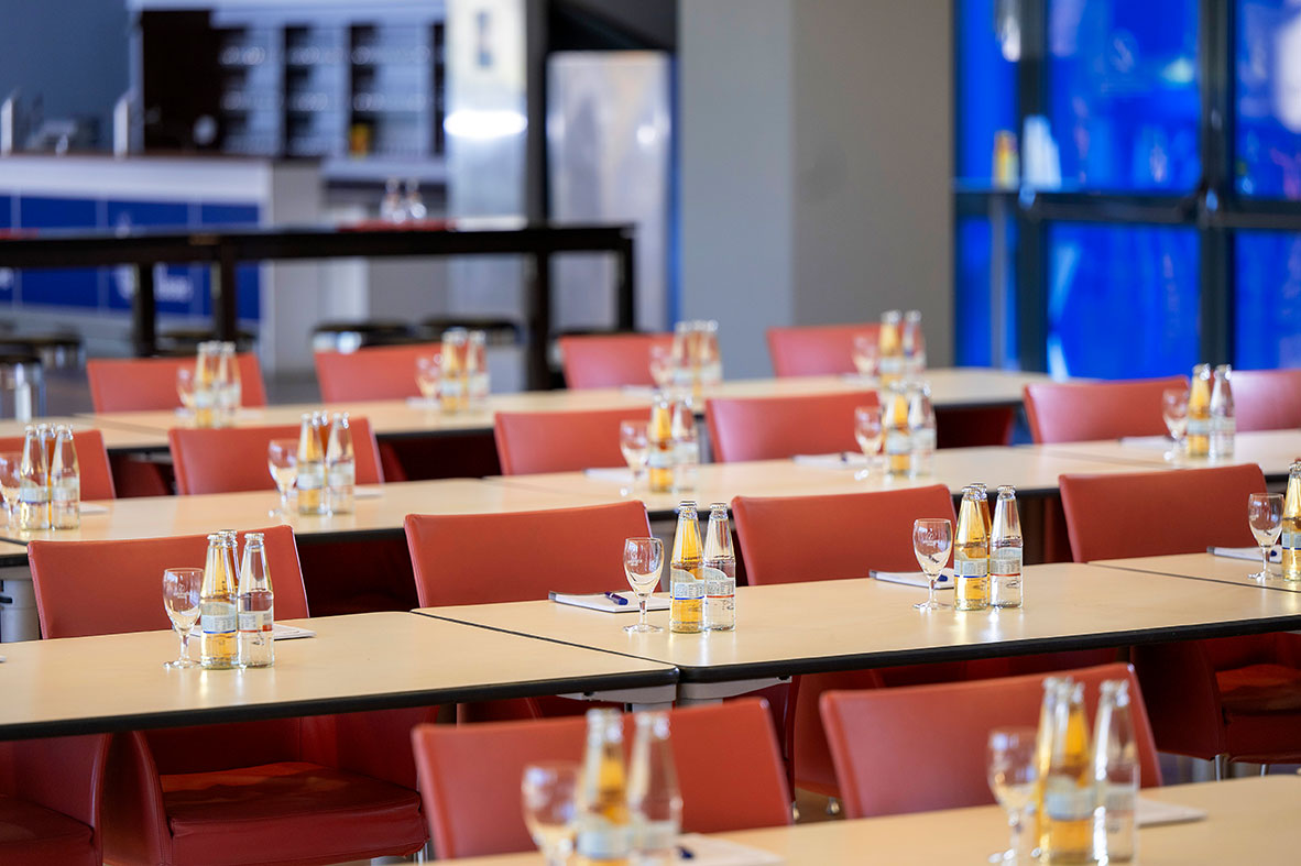 Ein großer Veranstaltungsraum mit Tischreihen und roten Stühlen. Die Tische sind mit Getränken und Notizblöcken eingedeckt.