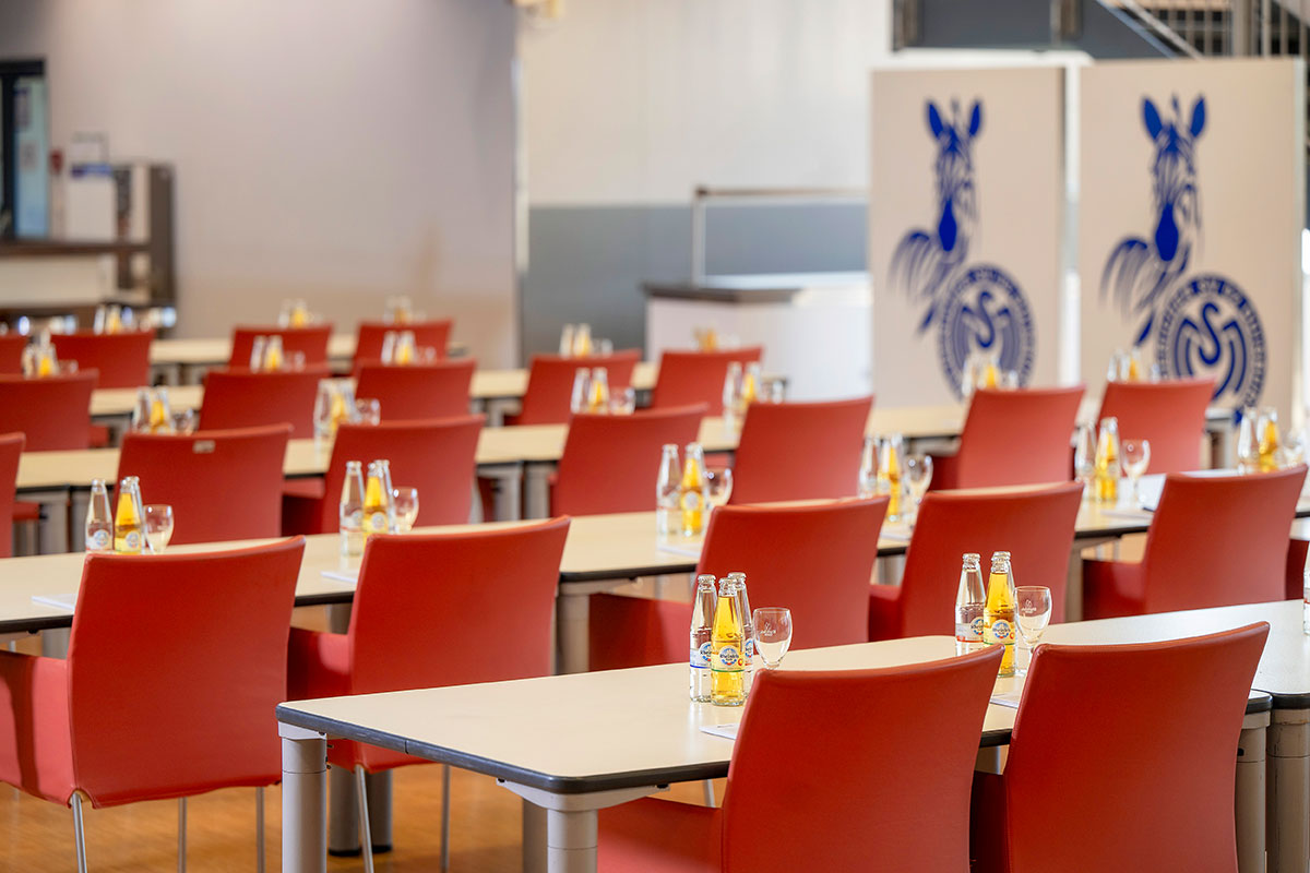 Ein großer Veranstaltungsraum mit Tischreihen und roten Stühlen. Im Hintergrund stehen MSV-Zebralogos