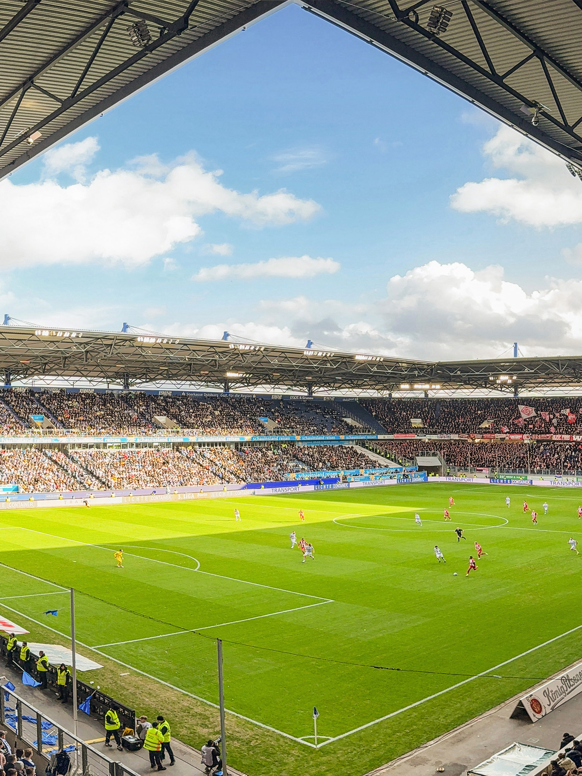 Ein Fußballstadion mit vielen Zuschauern und einem Spiel auf dem grünen Rasen unter blauem Himmel.