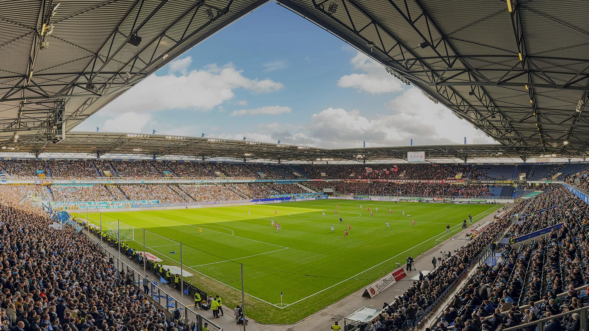 Ein Fußballstadion mit vielen Zuschauern und einem Spiel auf dem grünen Rasen unter blauem Himmel.