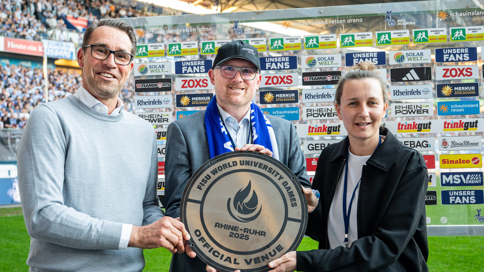 (v.l.): Michael Preetz, Sören Link, Sina Diekmann / Foto: Stadt Duisburg Zwei Männer und eine Frau nehmen vor einer gläsernen Sponsorenwand mit Logos auf einem Rasen in einem Stadion eine Plakette entgegen.