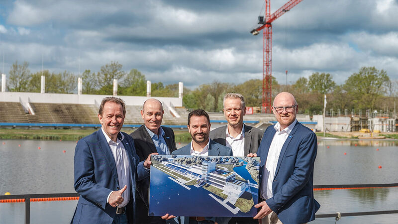 Fünf Männer im Anzug stehen vor den Regattabahn in Duisburg und halten ein Bild eines in den Händen.