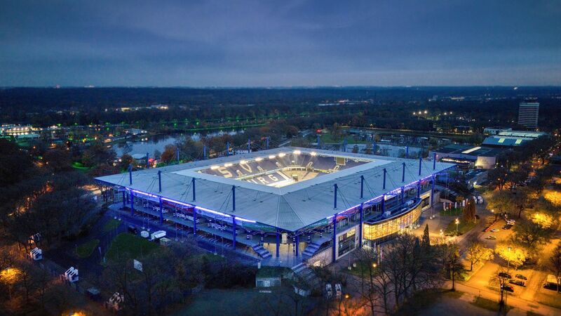 Luftaufnahme der beleuchteten Schauinsland-Reisen-Arena in Duisburg bei Nacht, umgeben von Bäumen.